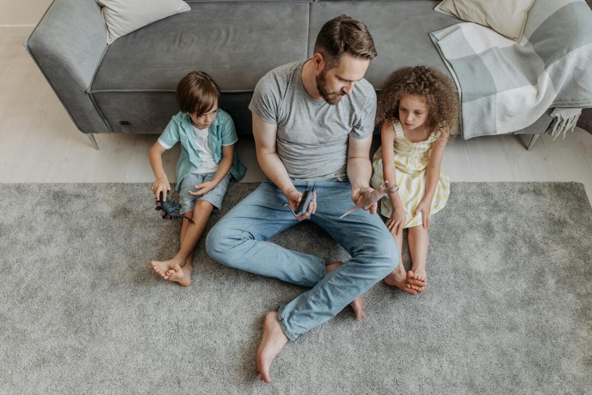 A Man in Gray Shirt Sitting on the Floor Between His Kids while Holding Toys, minimalism