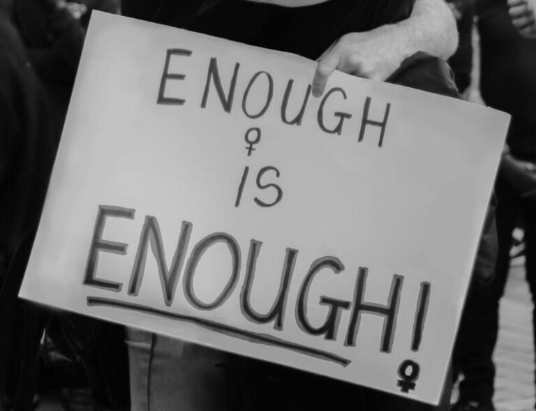 Black and white of unrecognizable couple hugging and standing on street with sign with ENOUGH IS ENOUGH inscription against crowd of people