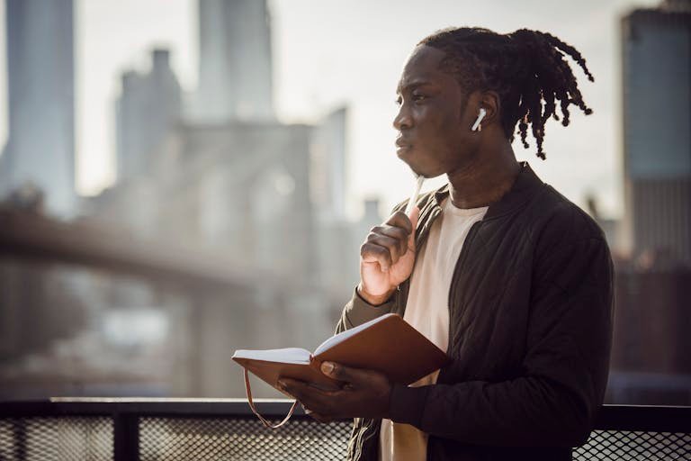 Thoughtful black man in wireless earphones with notepad on balcony - intentional living